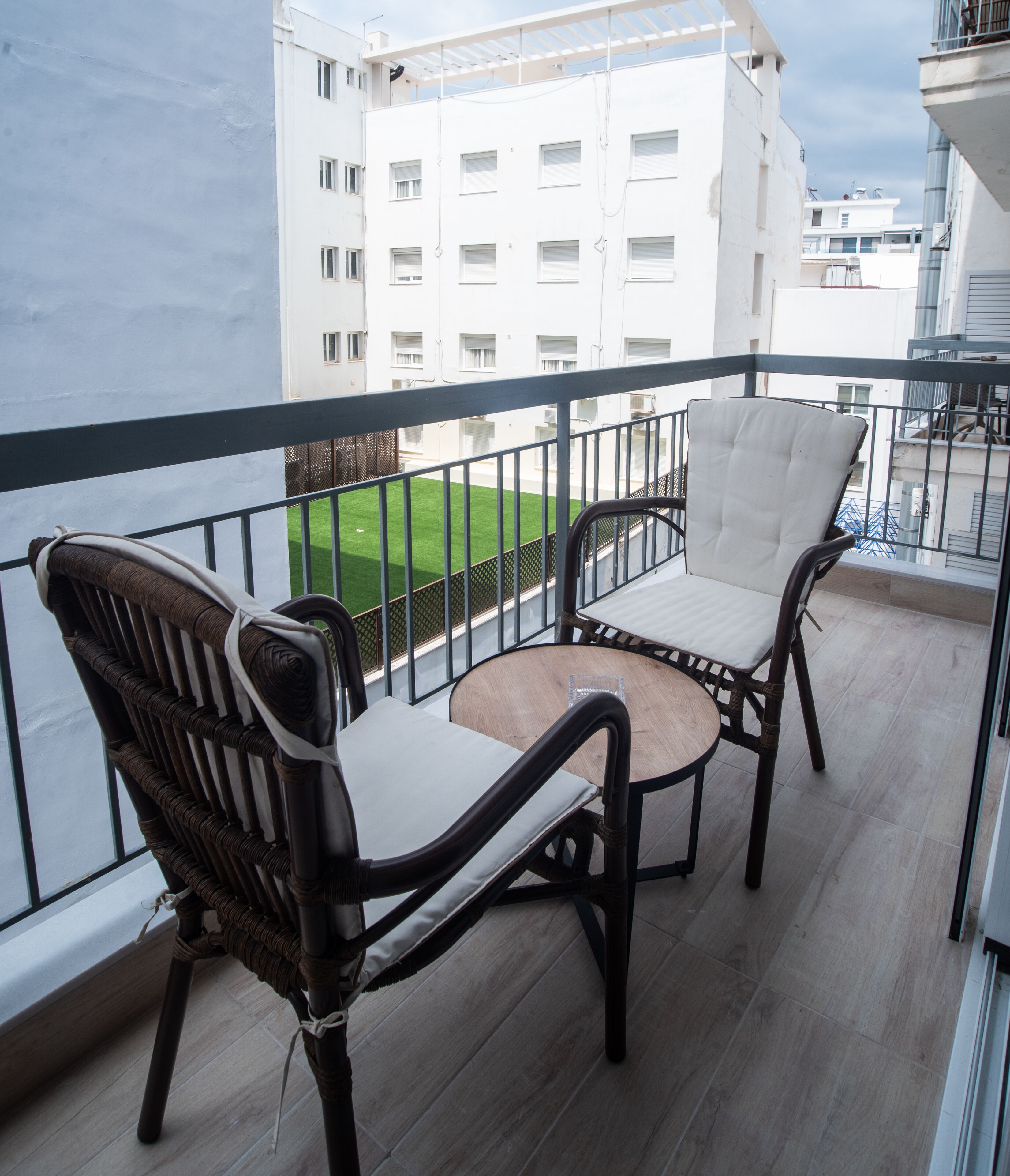 Un balcón con sillas de mimbre y una pequeña mesa, con vista a un área verde y edificios.