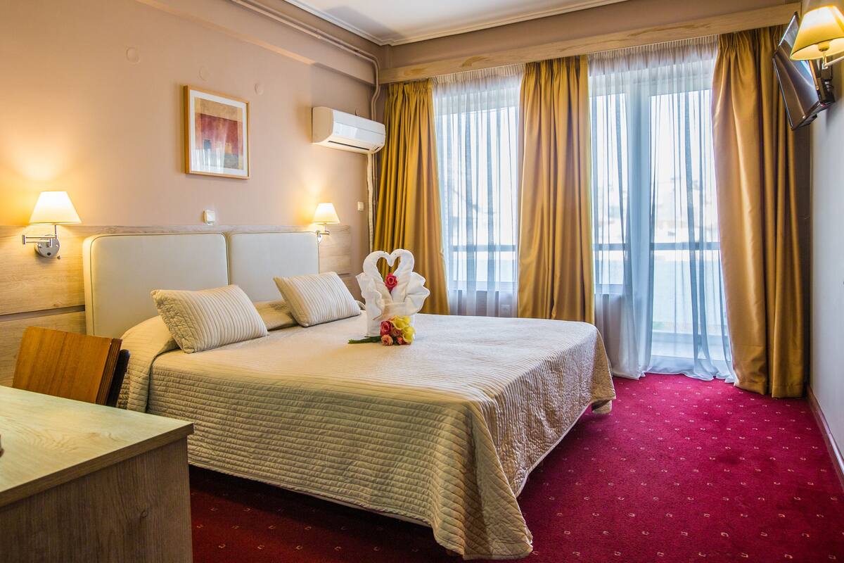A hotel room with a bed, bedside lamps, a desk, and a window with curtains.