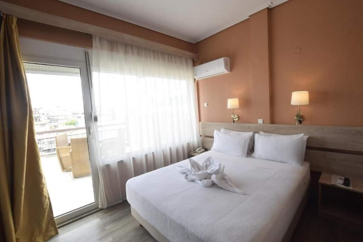 A hotel room with a large bed, a sliding glass door to the balcony, a wall-mounted air conditioner, and two bedside lamps.