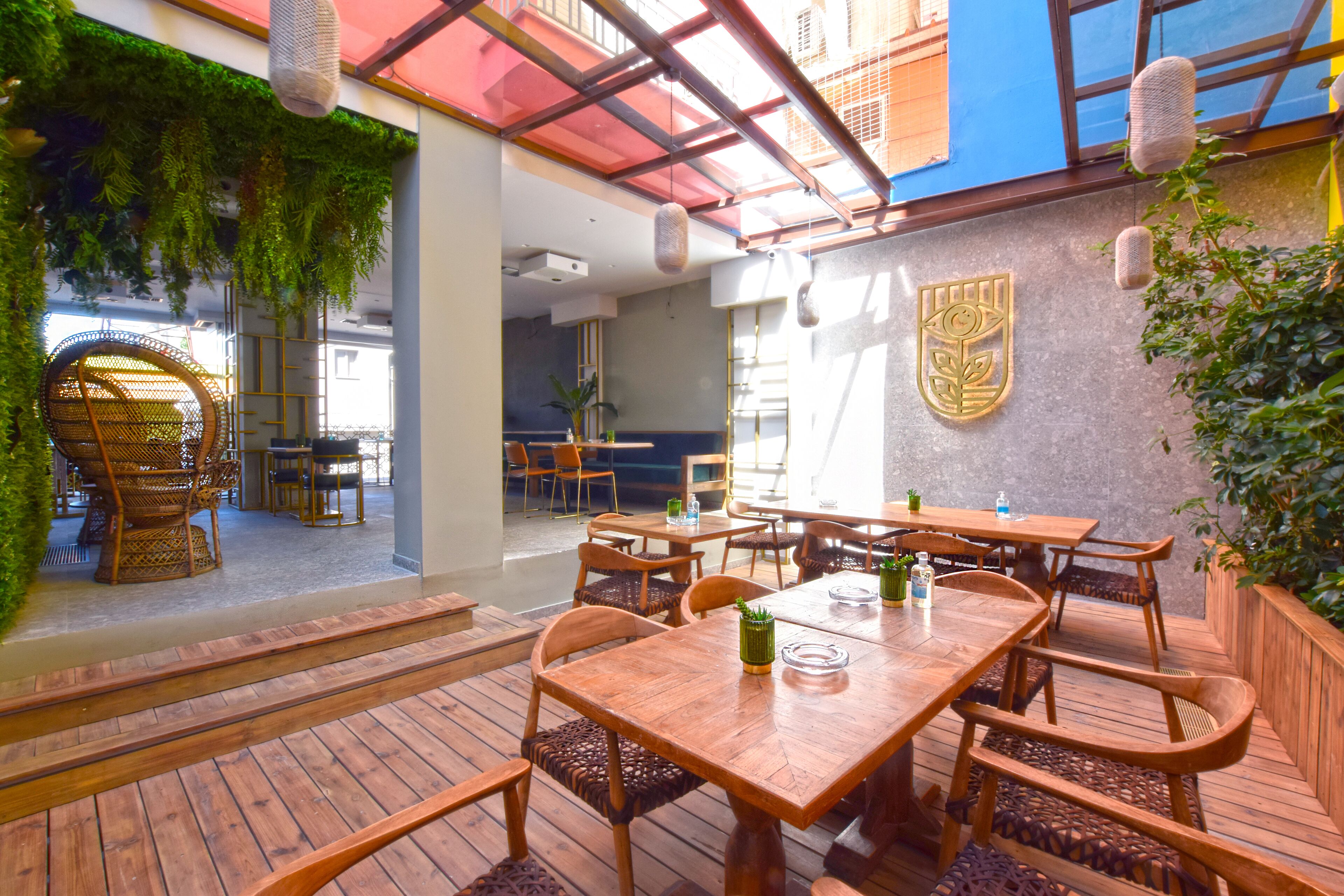 An indoor dining area with wooden tables and chairs, a green wall with hanging plants, and a view of the street outside.