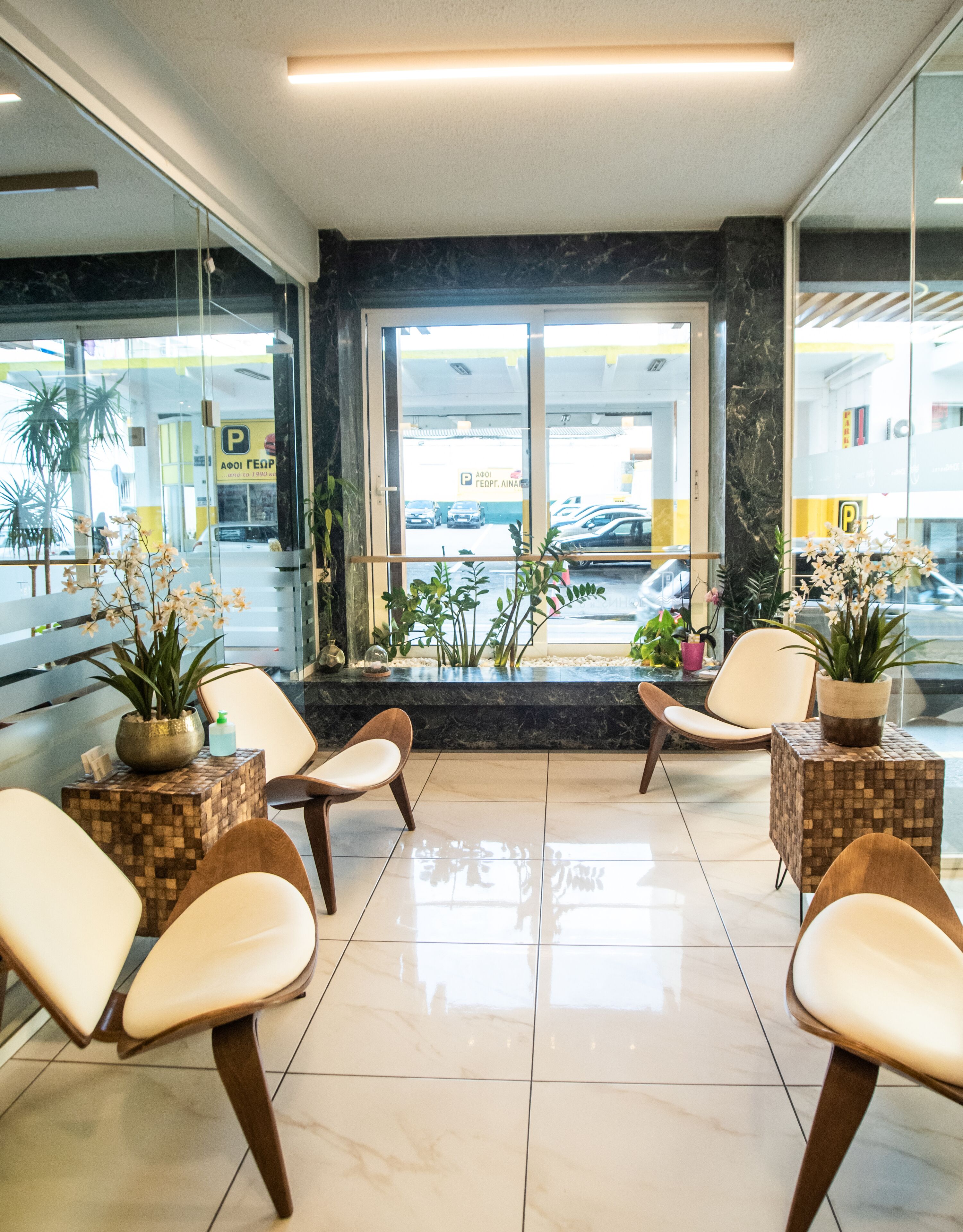 A reception area with a marble floor, seating with cushions, potted plants, and a view of parked cars outside.
