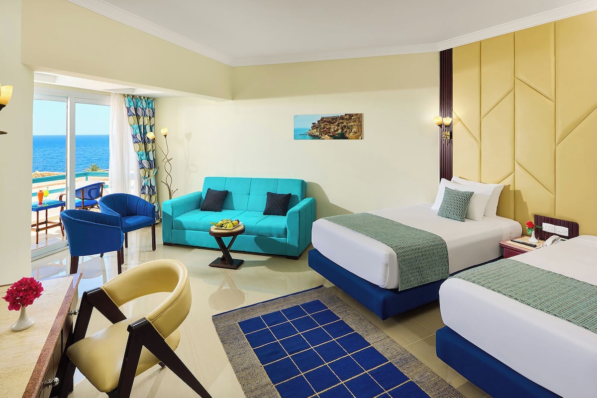 A hotel room with two beds, a sofa, a chair, a small table, and a view of the sea.