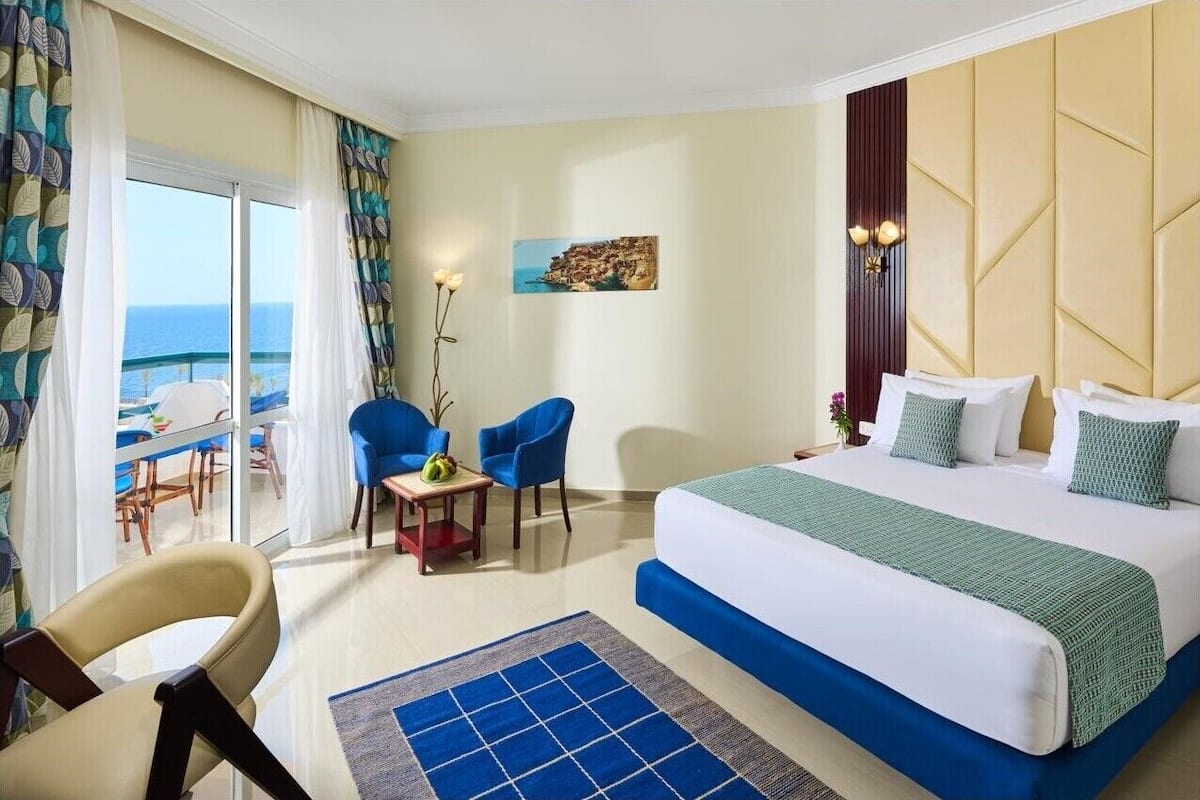 A hotel room with a bed, two chairs, a small table, and a view of the sea.