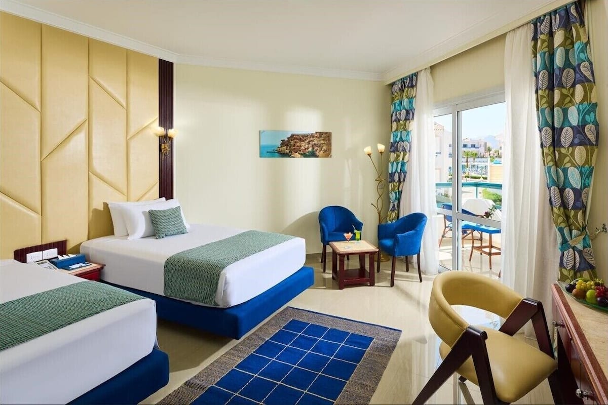 A hotel room with two beds, a blue armchair, a small table, and a view of the outside.