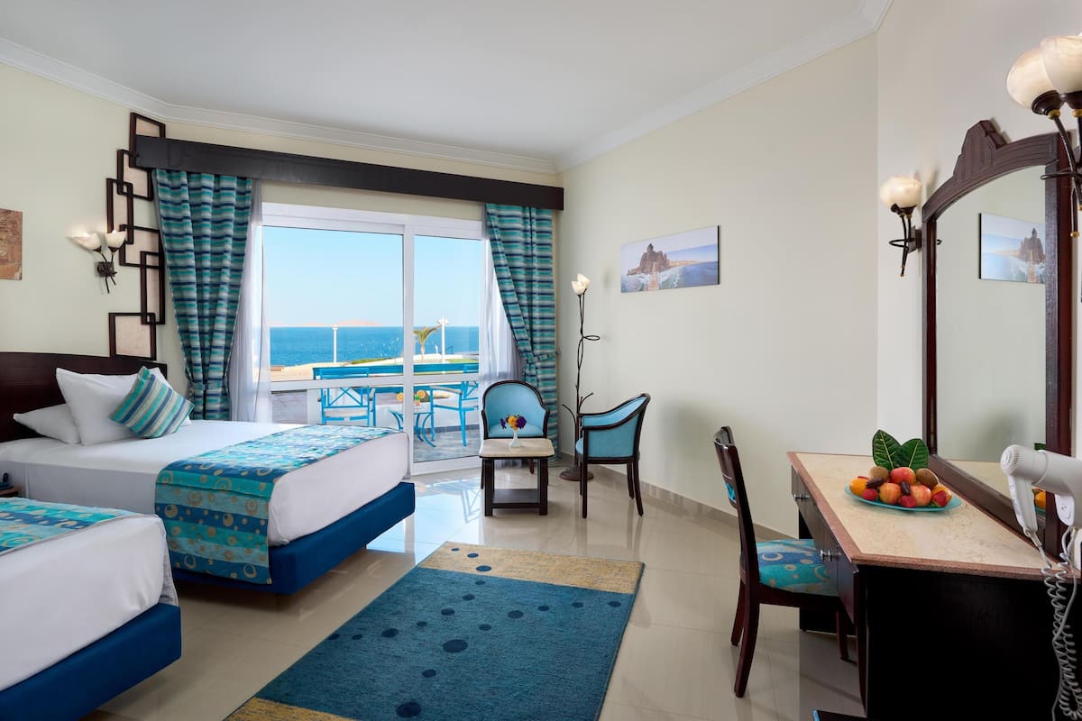 A hotel room with two beds, a dining table with fruit, a chair, a desk with a lamp, and a view of the ocean.