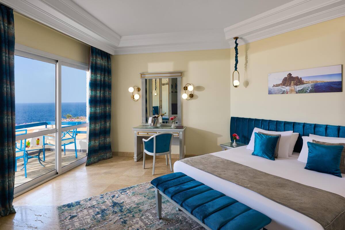 A hotel room with a large bed, a desk, a chair, and a view of the sea through a sliding door.