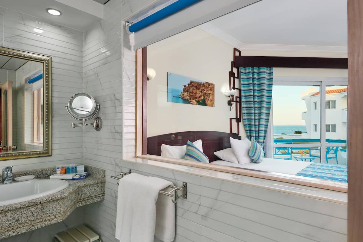 A hotel bathroom with a marble vanity, a large mirror, a round mirror, a towel rack, and a view of a balcony with a bed and a painting.