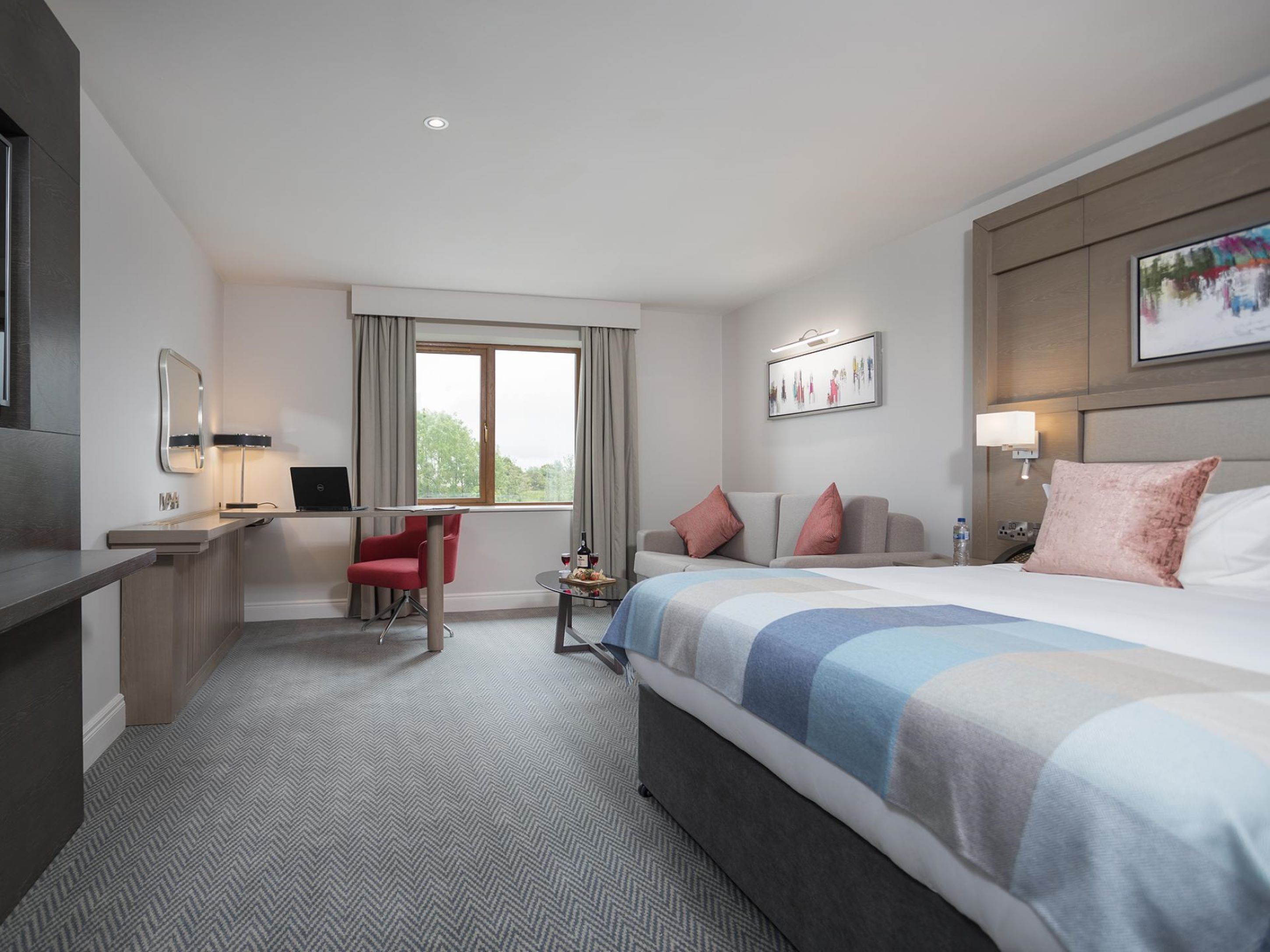 Ein modernes Hotelzimmer mit einem großen Bett, einem Schreibtisch mit einem Laptop, einem roten Sessel, einem kleinen Tisch mit einer Flasche und Gläsern sowie einem Fenster mit Vorhängen.