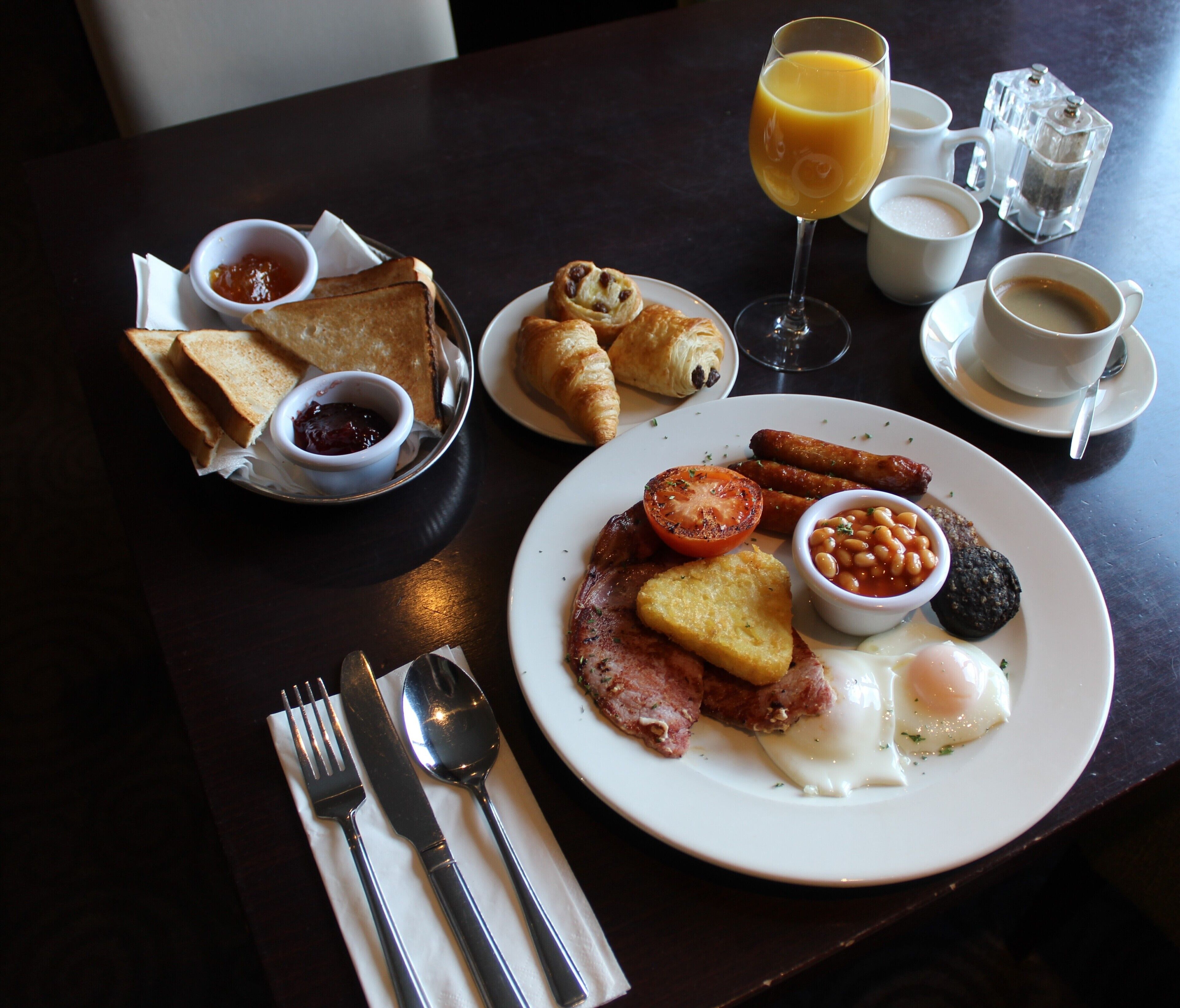 A breakfast table with toast, croissants, sausages, eggs, tomatoes, beans, and a glass of orange juice.