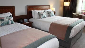 A hotel room with two beds, each with a wooden headboard, white linens, and matching throw pillows. There is a bedside table with a lamp and a phone on one side, and a small table with a wine glass and a bottle on the other.