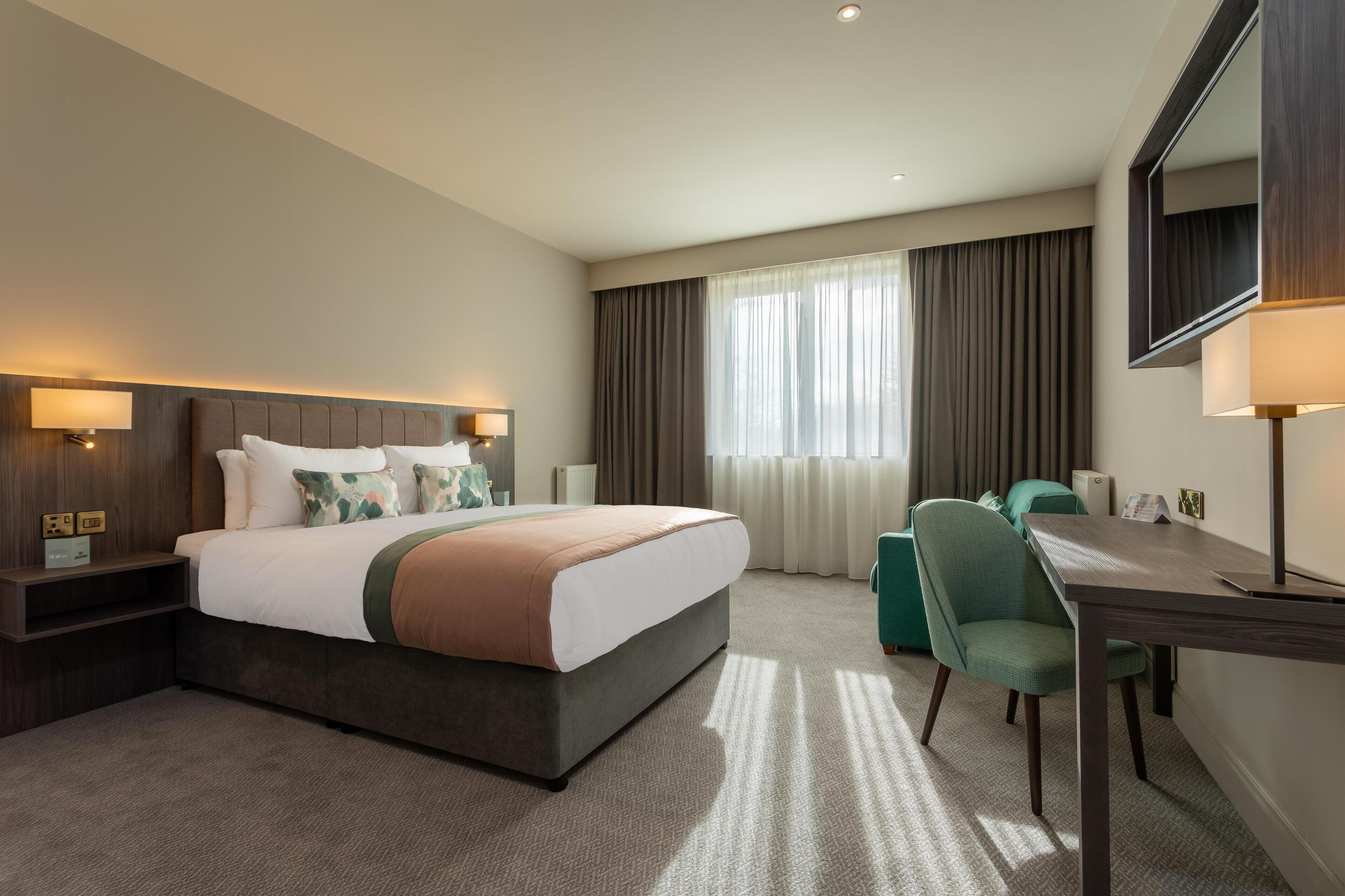 Una habitación de hotel con una cama grande, un escritorio, una silla y un ventanal con cortinas.