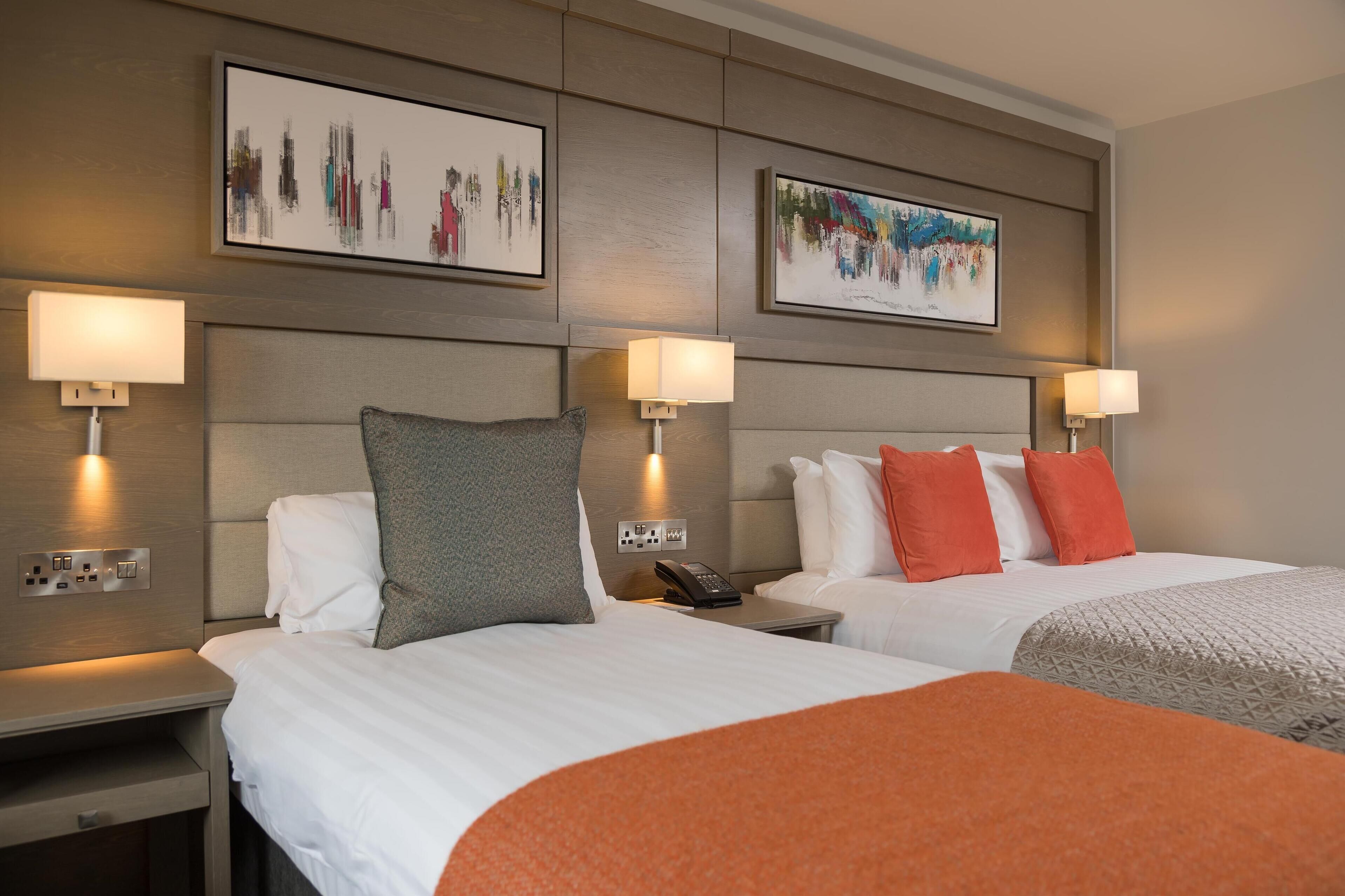 Habitación de hotel con dos camas, cada una con almohada y manta. Hay lámparas de pared a cada lado de las camas.