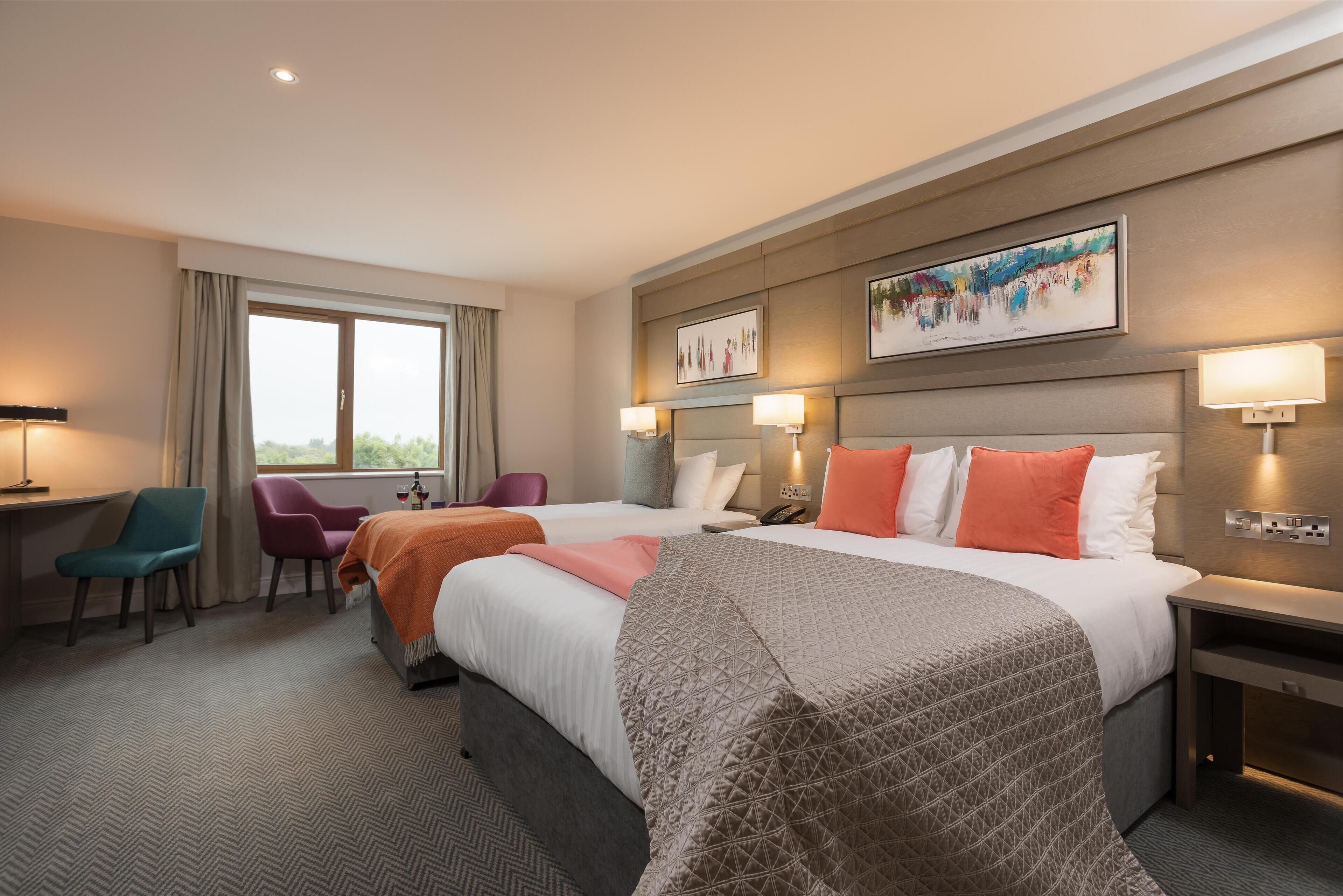 Habitación de hotel con una cama grande, dos mesitas de noche, un escritorio y una silla. Hay dos ventanas con cortinas, un cuadro en la pared y una lámpara en cada mesita de noche.