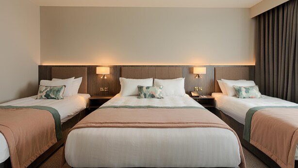 Habitación de hotel con tres camas dispuestas en fila, cada una con cabecera, mesitas de noche y lámparas.