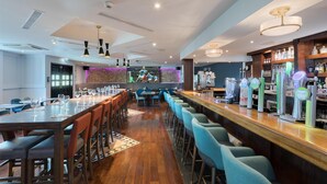 Een moderne bar met een lange toonbank, barstoelen en een televisiescherm waarop sport wordt vertoond.