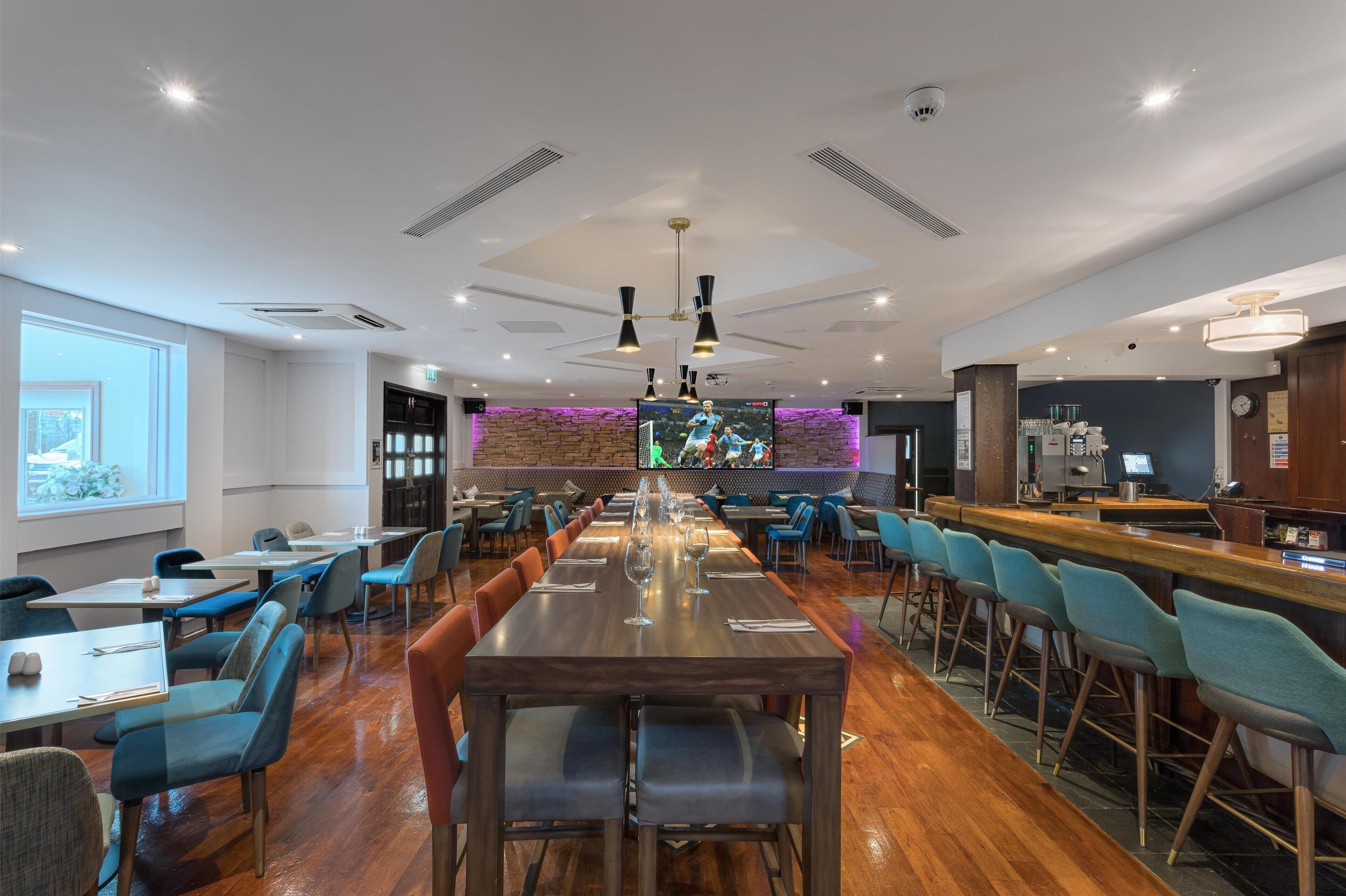 Een ruim restaurant met lange gedeelde tafels en zitplaatsen aan de bar, een groot scherm om naar te kijken, en een modern interieur.