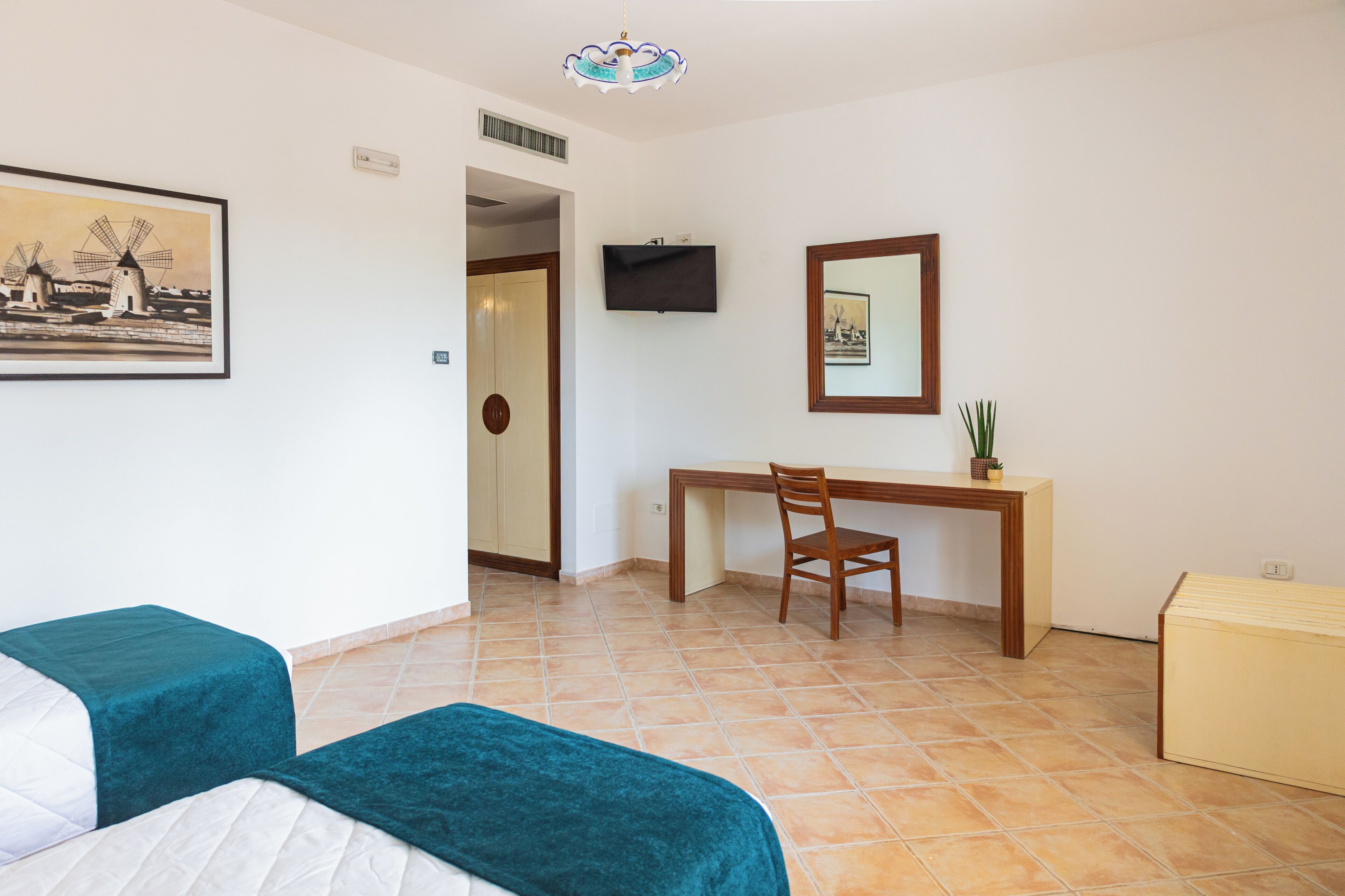 Quarto de hotel com cama, escrivaninha, cadeira, televisão e um quadro.