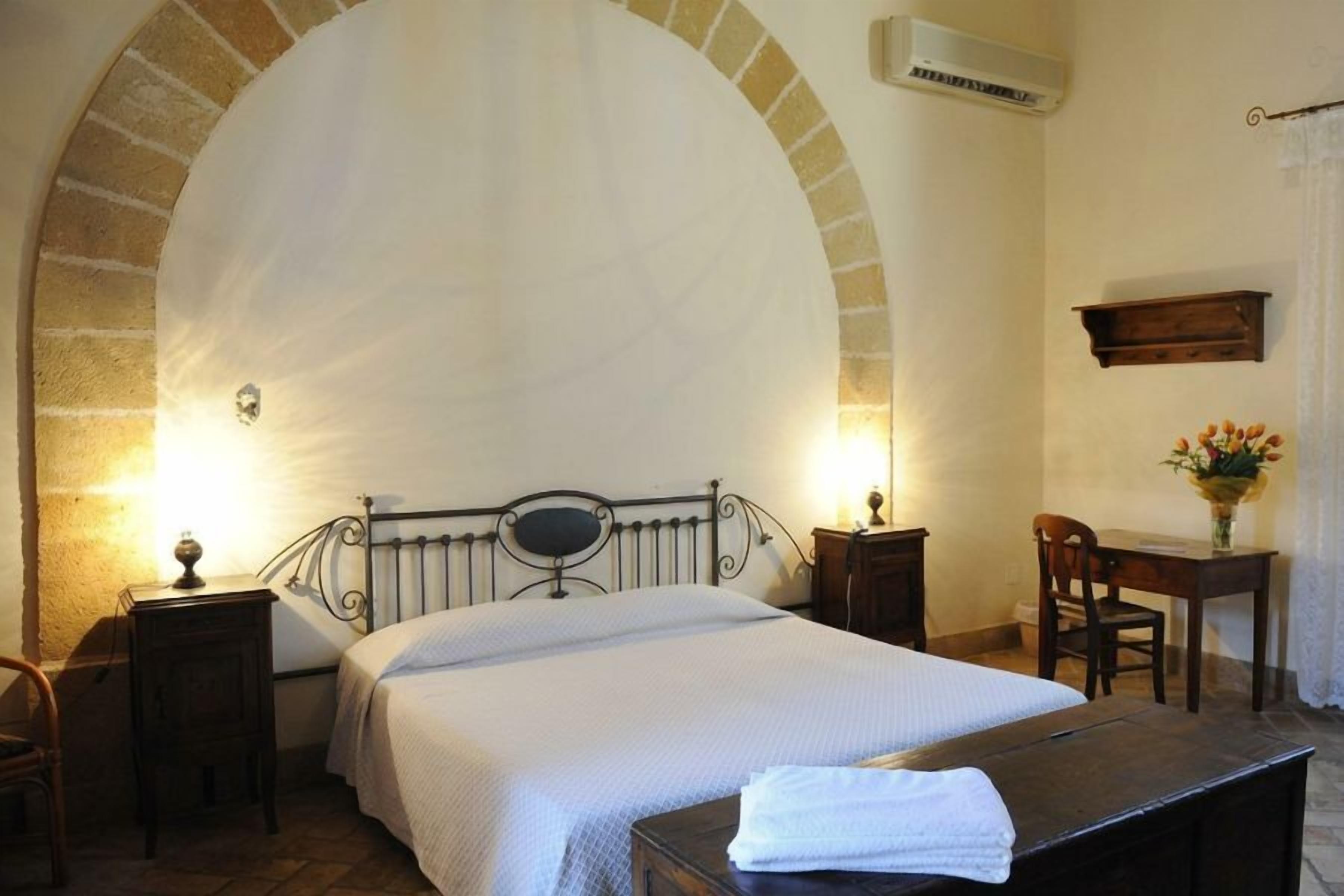 Quarto com uma grande janela em arco, cama com colcha branca, mesa de cabeceira com abajur, cadeira de madeira e pequena escrivaninha com um vaso de flores.