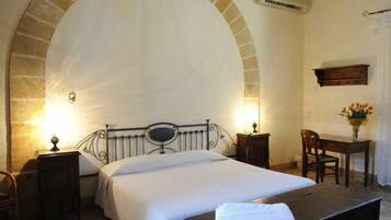 Quarto com uma grande janela em arco, cama com colcha branca, mesa de cabeceira com abajur, cadeira de madeira e pequena escrivaninha com um vaso de flores.