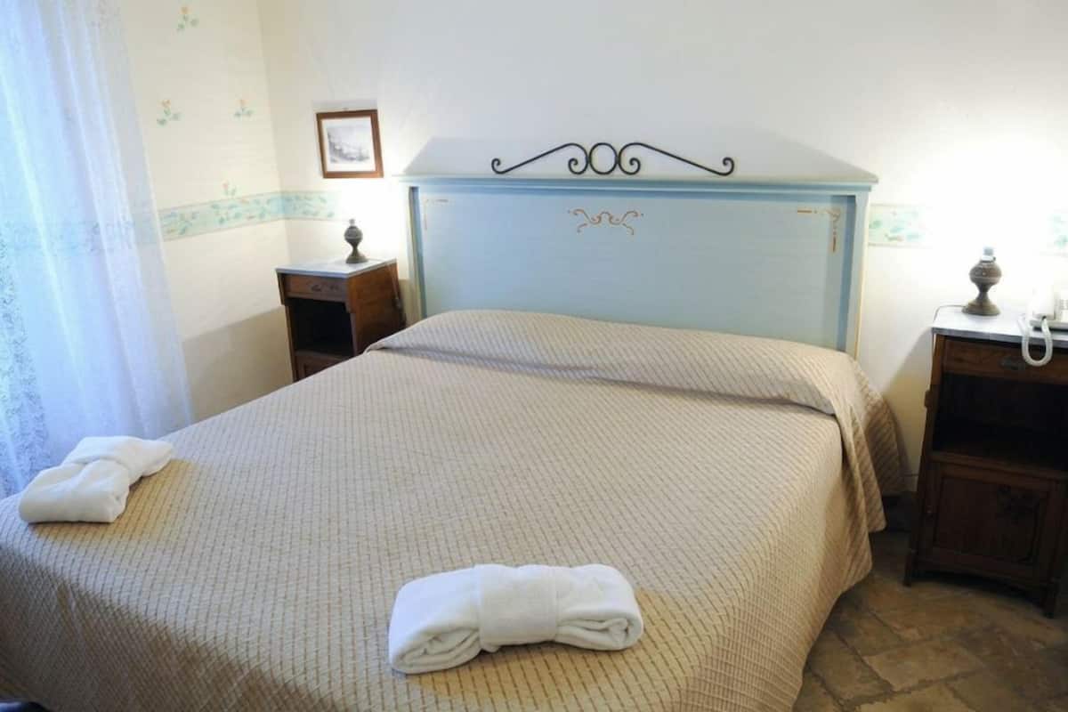 A neatly made bed with a headboard, a bedside table with a lamp, and a wall with a floral pattern.