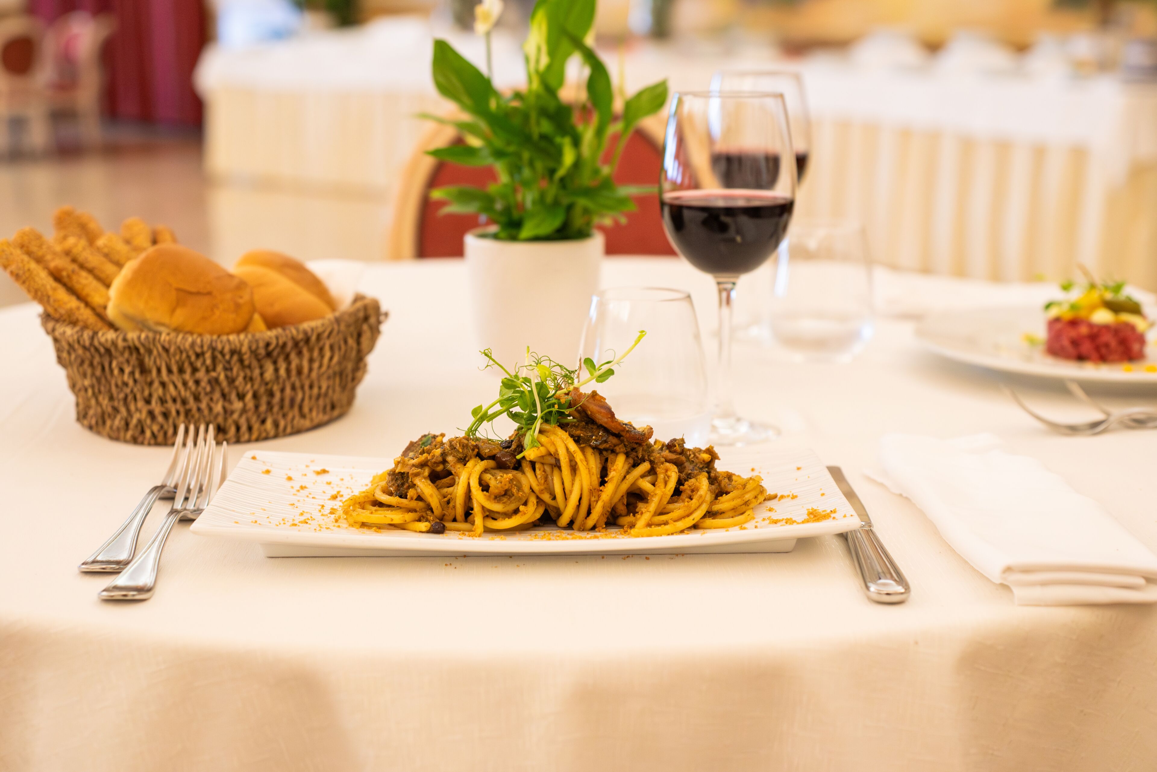 Une table à manger avec une assiette de spaghettis, un verre de vin rouge, un panier de pain et une plante en pot.