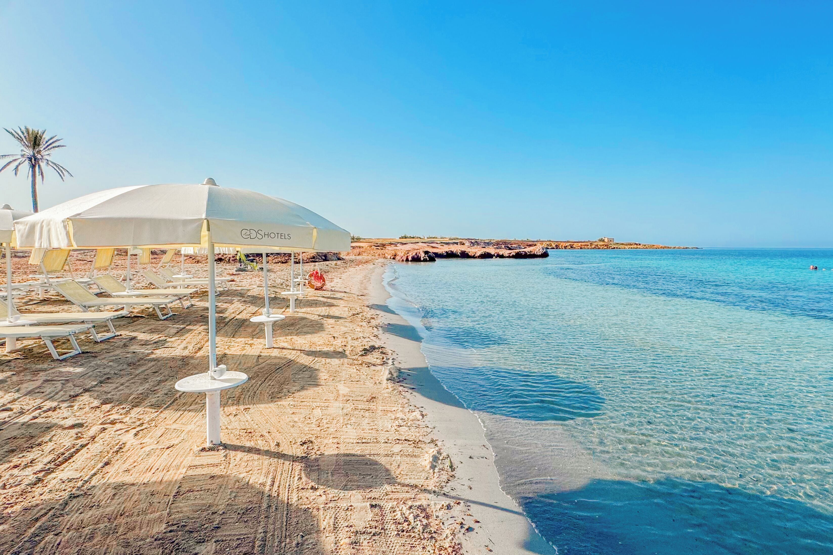 Een hotel aan het strand met ligbedden en parasols, een heldere blauwe zee en een rotsachtige kustlijn.