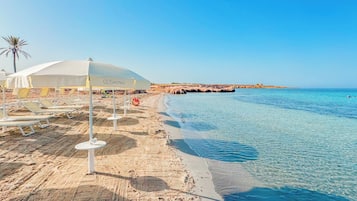 Een hotel aan het strand met ligbedden en parasols, een heldere blauwe zee en een rotsachtige kustlijn.