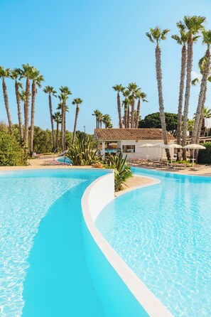 A swimming pool with a curved edge, surrounded by palm trees and a building with a tiled roof. - Baglio Basile Hotel (Petrosino)