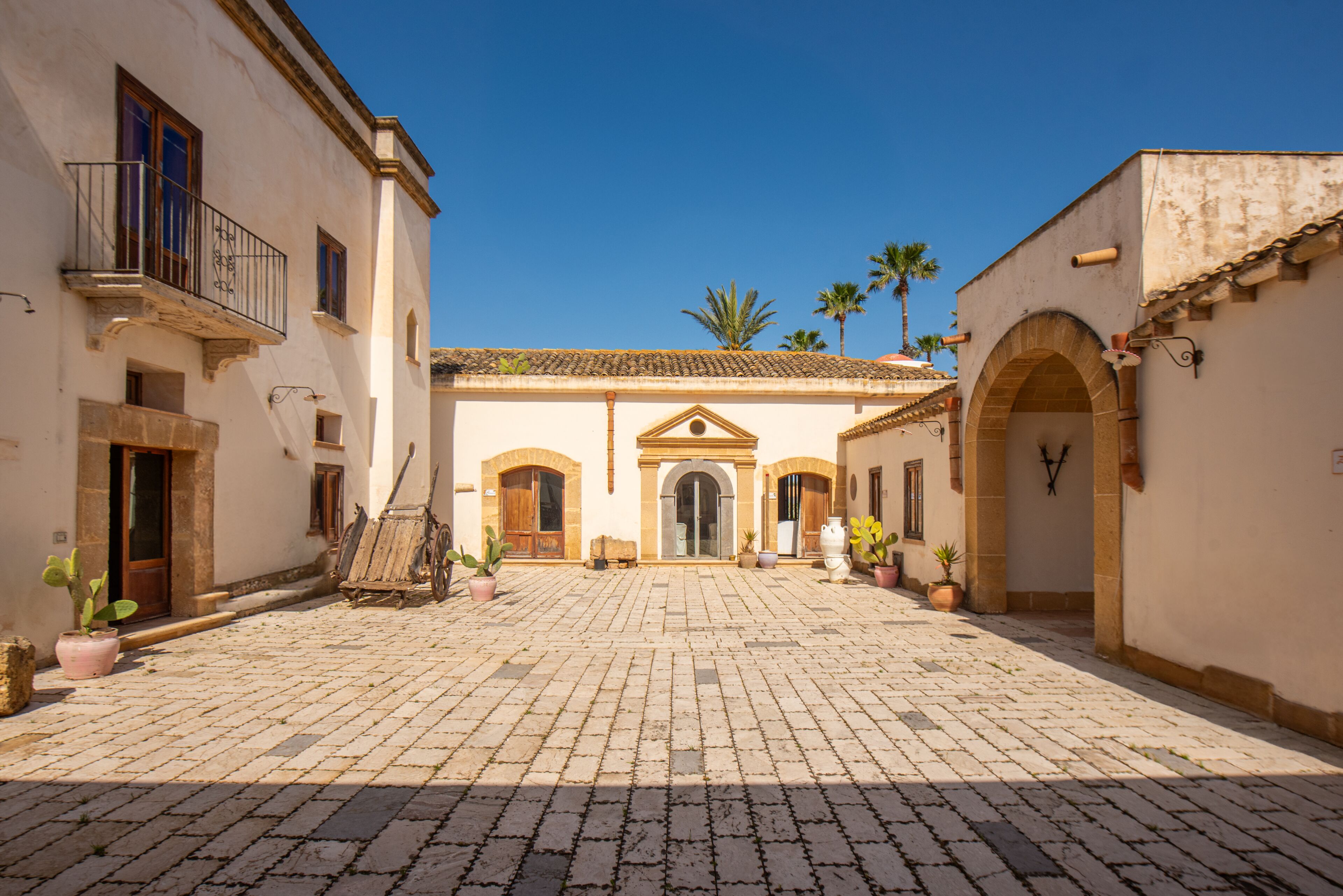 A courtyard with cobblestone paving, a central building with an arched entrance, and potted plants.