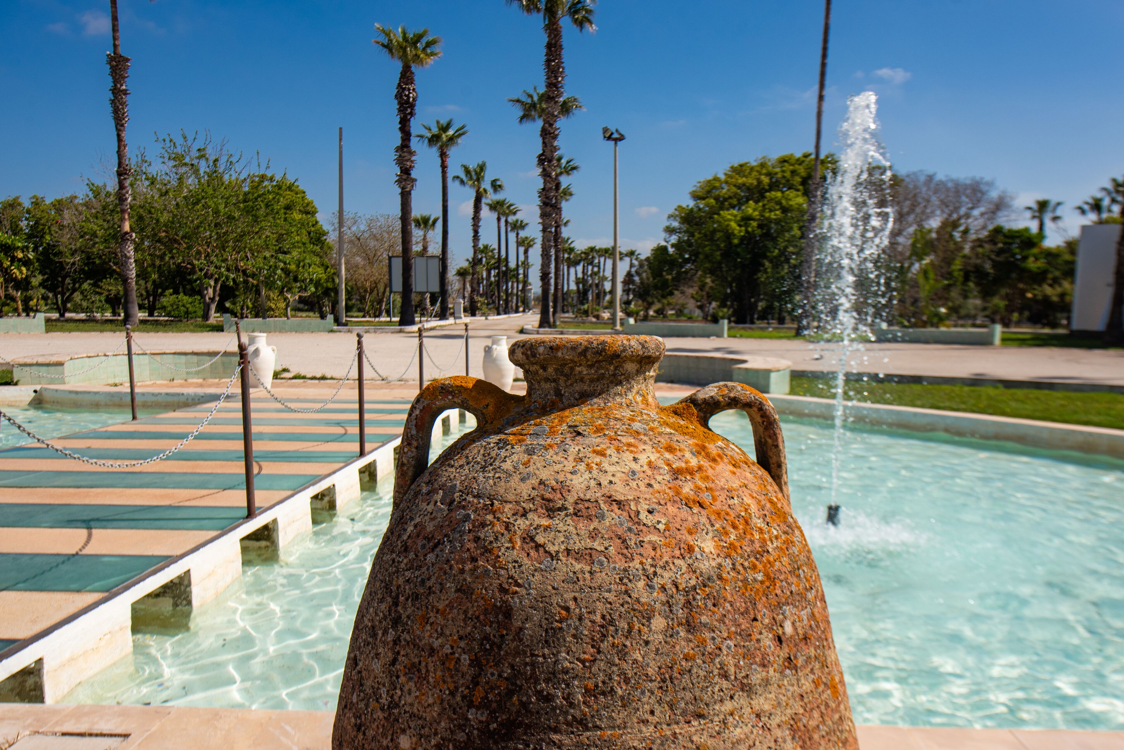 Un'ampia brocca di ceramica arrugginita con manici, posizionata in una piccola vasca con una fontana sullo sfondo, circondata da palme e un cielo sereno.