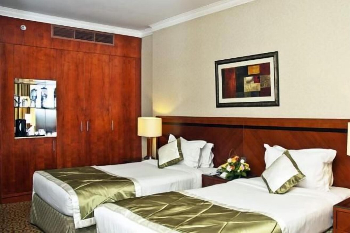 Habitación de hotel con dos camas, un armario de madera, un cuadro enmarcado en la pared y una mesita de noche con una lámpara.