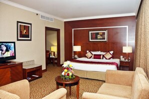A hotel room with a bed, two bedside lamps, a TV, a wooden desk, a chair, a small table with flowers, and a carpeted floor.