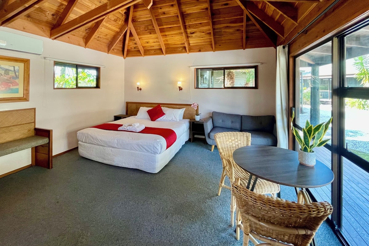 Ein Schlafzimmer mit Holzbalkendecke, einem Bett mit weißer Bettwäsche und roten Kissen, einem grauen Sofa, einem Korbsessel, einem kleinen Tisch mit Topfpflanze und einem Fenster mit Blick auf Grünflächen.