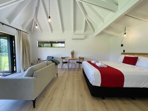 A modern hotel room with a large bed, a sofa, a dining table, and a view of the outdoors. - Pacific Harbour Villas (Tairua)