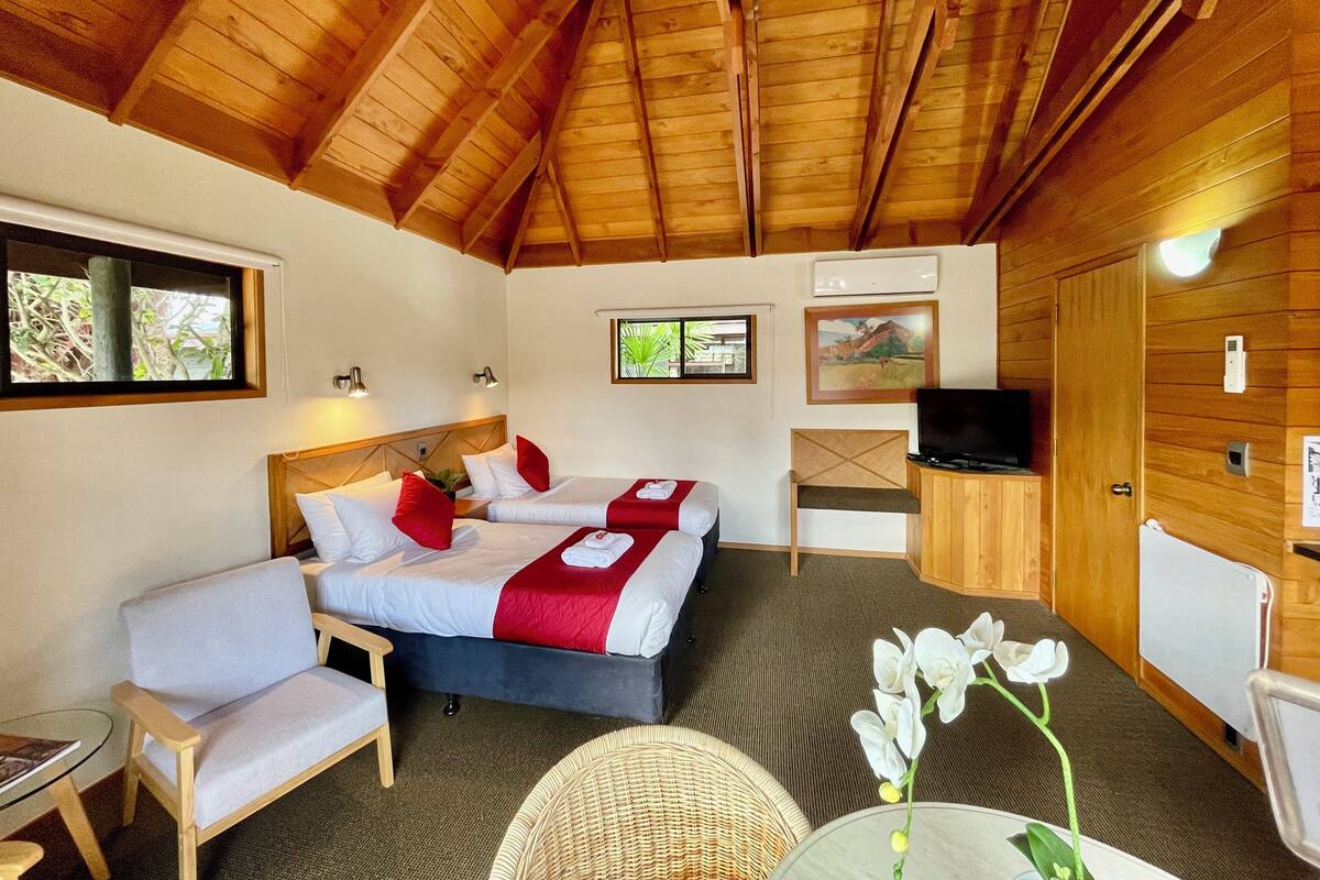 Ein Zimmer mit Holzbalkendecke, zwei Betten mit rot-weißer Bettwäsche, einem Fernseher, einem Schreibtisch, einem Stuhl und einem kleinen Tisch mit einer Blume.