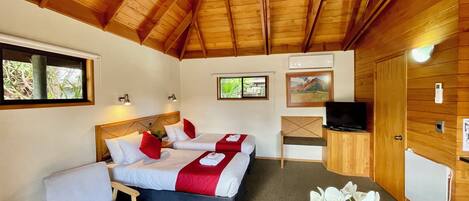 A room with a wooden ceiling, two beds with red and white bedding, a TV, a desk, a chair, and a small table with a flower.