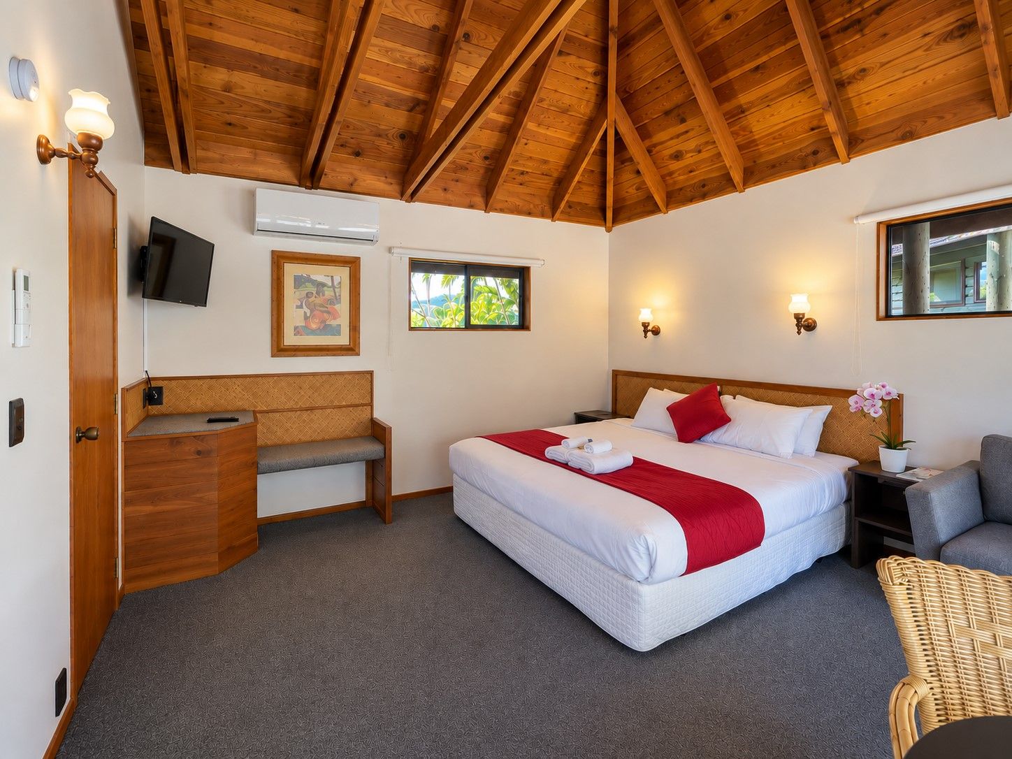 A hotel room with a wooden ceiling, a bed with white bedding and red accents, a desk with a chair, a television, and a window with a view.