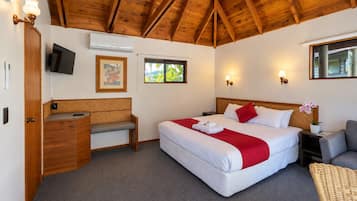 A hotel room with a wooden ceiling, a bed with white bedding and red accents, a desk with a chair, a television, and a window with a view.