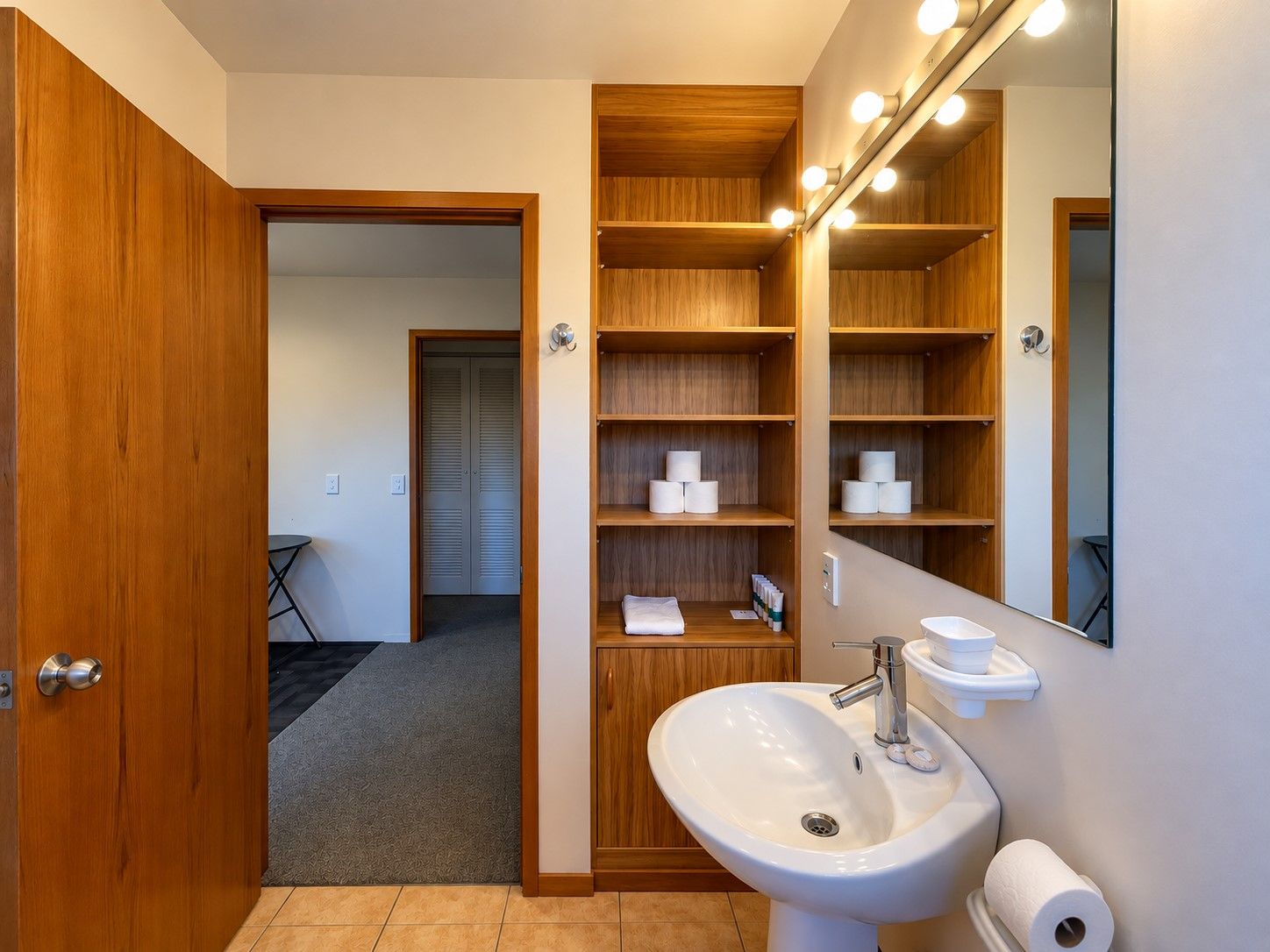 A bathroom with a wooden vanity, a sink, and a mirror with lights. There are shelves with toiletries and towels.