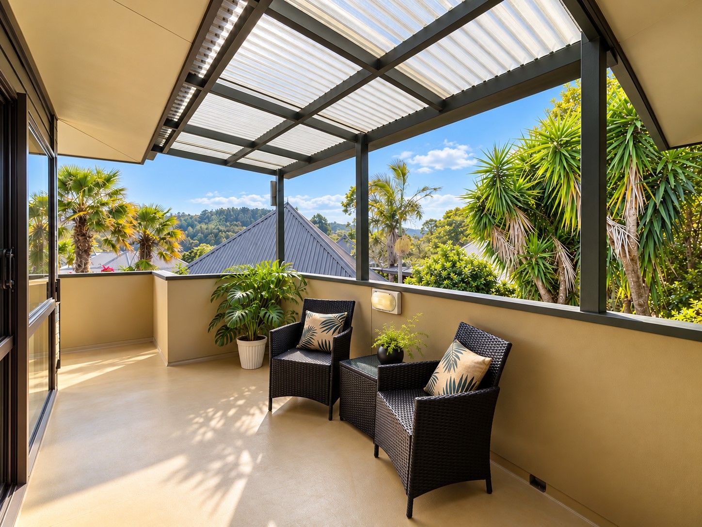 A covered balcony with two wicker chairs and a potted plant, overlooking a tropical landscape with palm trees and a mountain in the distance.
