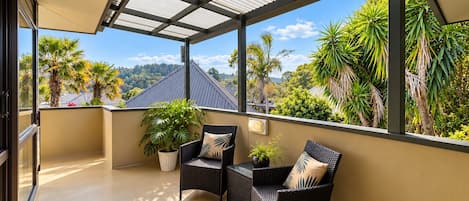 A covered balcony with two wicker chairs and a potted plant, overlooking a tropical landscape with palm trees and a mountain in the distance.