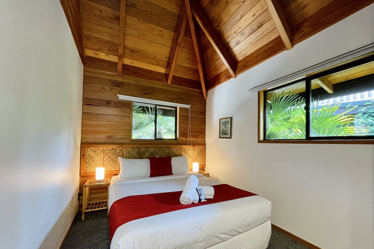 Eine Holzbalkendecke, ein Bett mit weißer und roter Bettwäsche, ein Nachttisch mit Lampe und ein Fenster mit Blick auf Grünflächen.