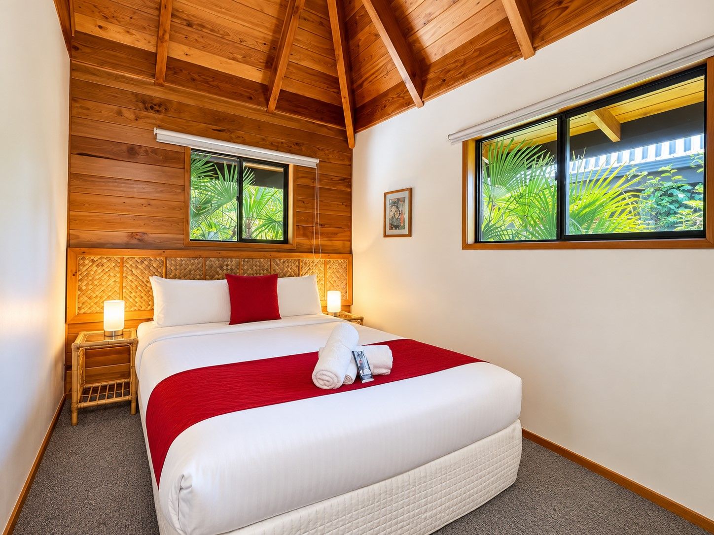 Une chambre d’hôtel avec un plafond en bois, un lit avec une literie blanche et un détail rouge, deux lampes de chevet et une fenêtre donnant sur un paysage verdoyant.