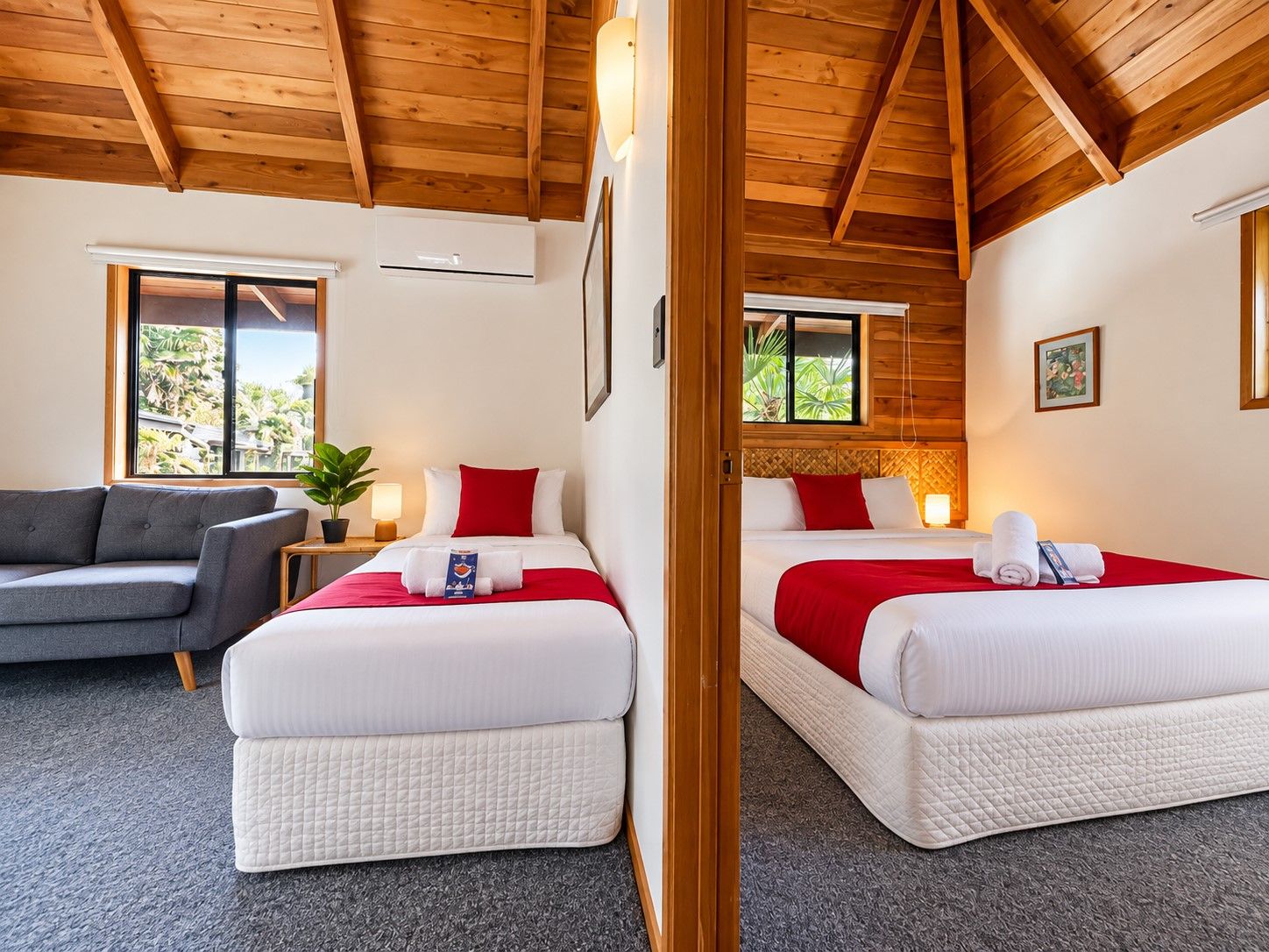 Habitación de hotel con dos camas, un sofá gris, una ventana con vistas y un techo de madera.