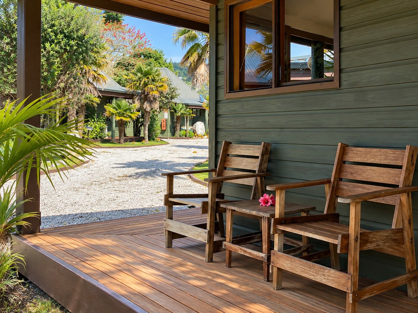 Een houten terras met twee stoelen en een tafel, met uitzicht op een grindpad met palmbomen en een gebouw op de achtergrond.