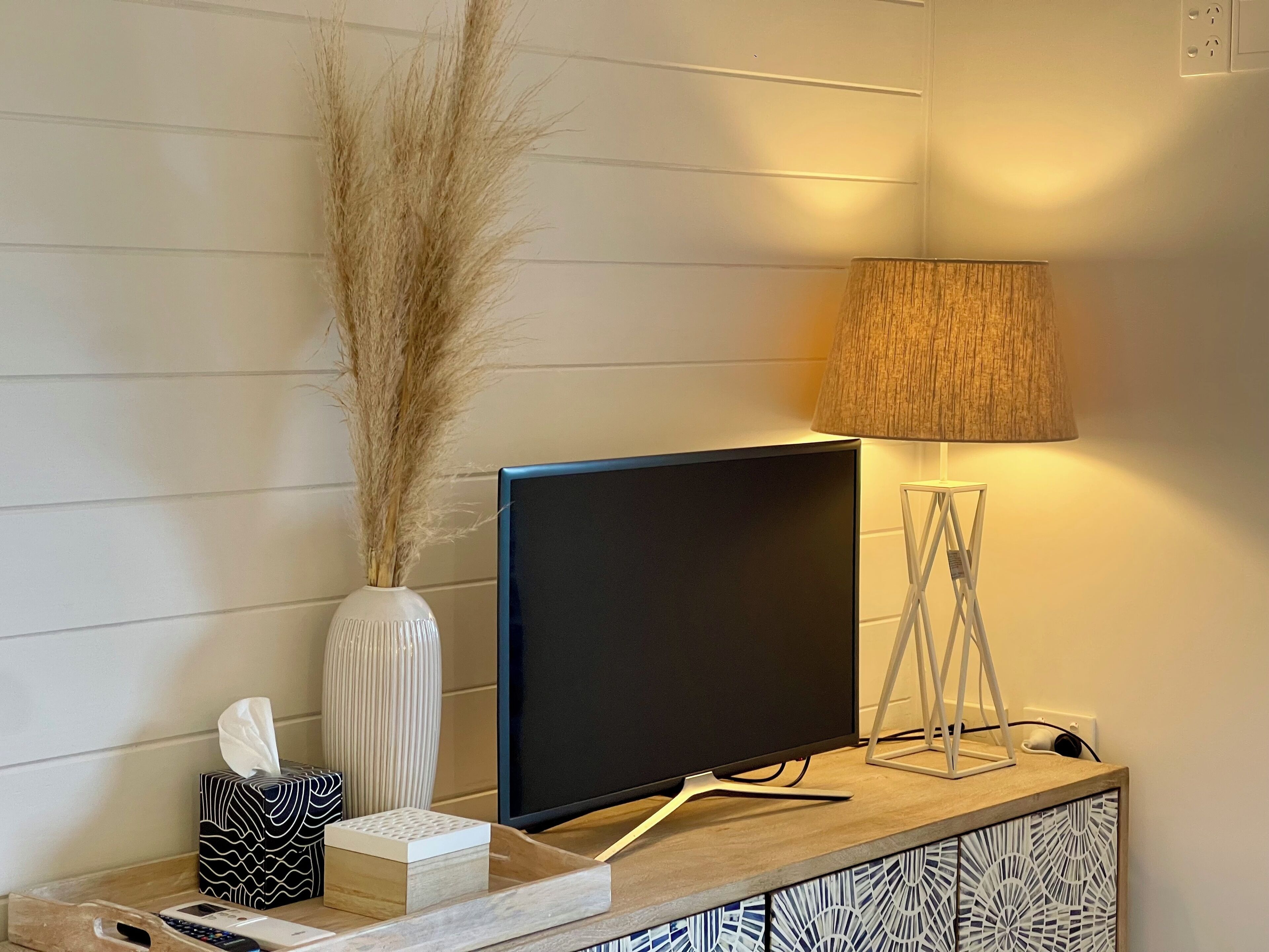 Ein modernes Wohnzimmer mit einem Flachbildfernseher auf einem Holzständer, einer Vase mit Trockenpflanzen, einer Lampe und einer Taschentuchbox.