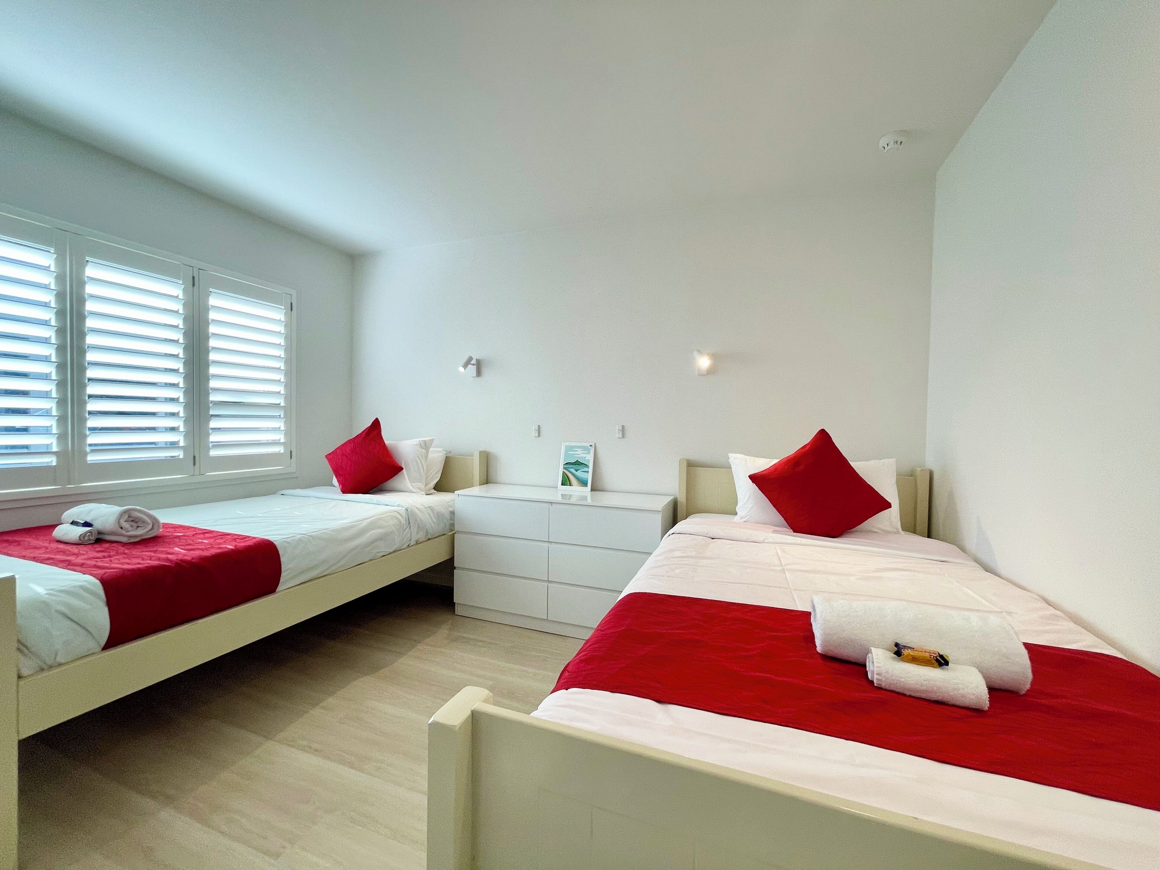 Een slaapkamer met twee bedden, elk voorzien van rode en witte beddengoed, een witte dressoir en een raam met jaloezieën.