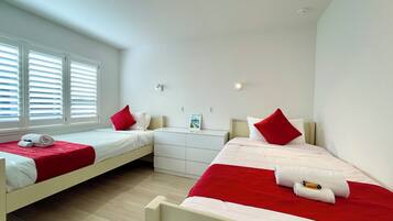 Een slaapkamer met twee bedden, elk voorzien van rode en witte beddengoed, een witte dressoir en een raam met jaloezieën.