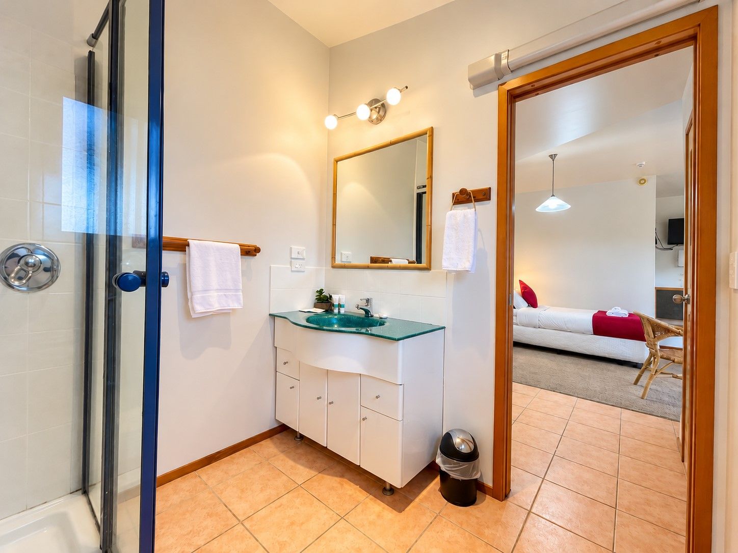 A bathroom with a glass shower enclosure, a white sink with a green countertop, a mirror above the sink, and a door leading to a bedroom with a bed and a red pillow.