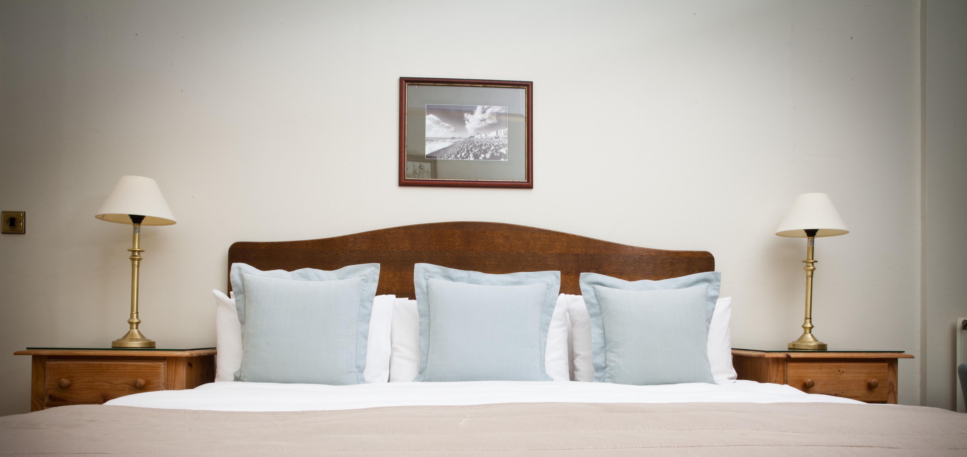 Een netjes opgemaakt bed met een houten hoofdbord, twee nachtkastjes met lampen en een ingelijst schilderij aan de muur.